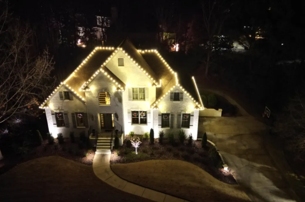 Aerial view of a white brick home with roofline lights and lit landscaping at night.
