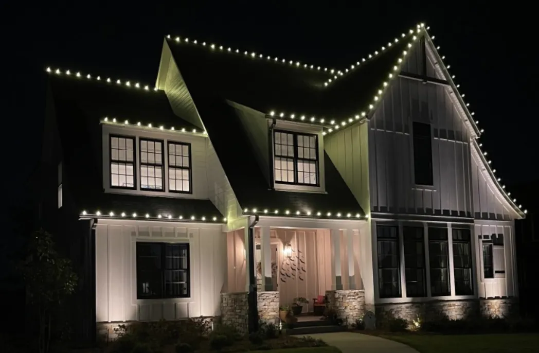 Roofline lights on a white board-and-batten home with three dormers at night.